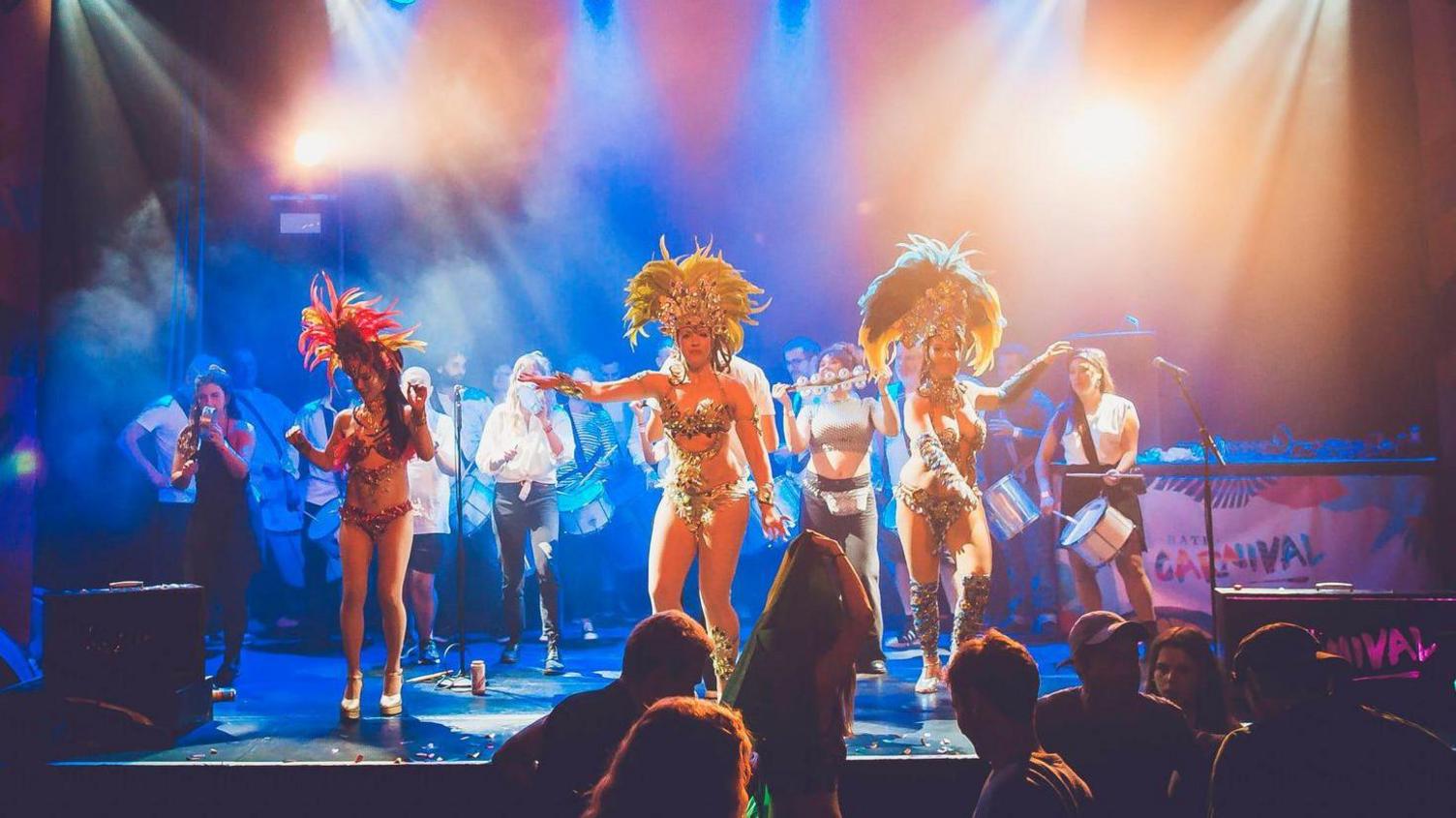 Musicians, playing drums, bells and a woodwind instrument, with carnival-style dancers in the foreground.