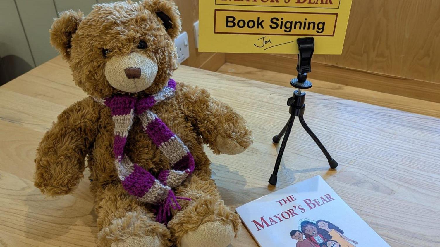 A teddy bear is sat on a table wearing a purple and white scarf. Next to him is a copy of The Mayor's Bear book.