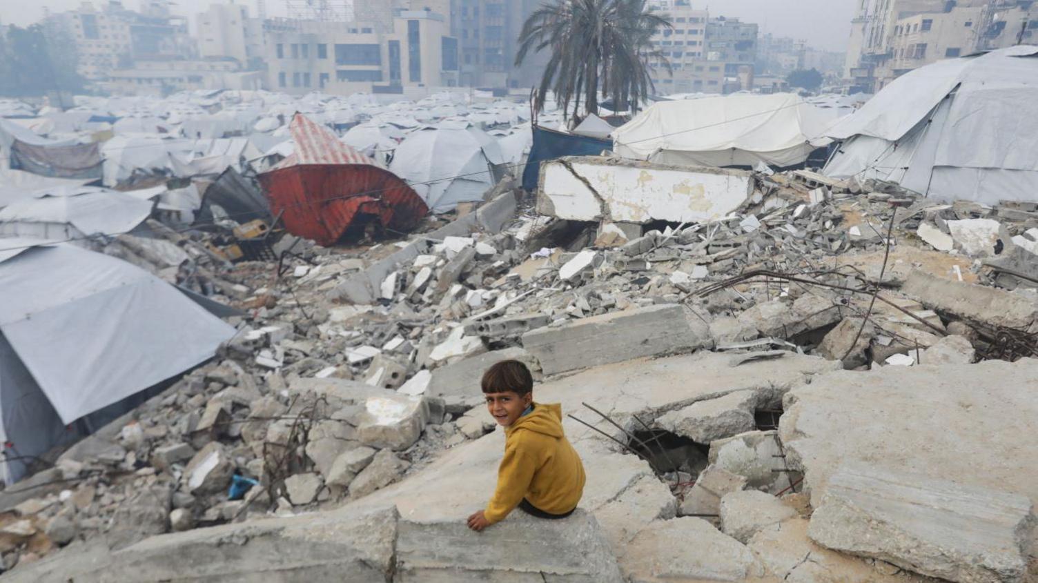 A Palestinian child wearing a yellow jumper sits amid the dusty grey rubble of buildings destroyed in previous Israeli strikes in Gaza City, with tents in the background, on 16 January 2025