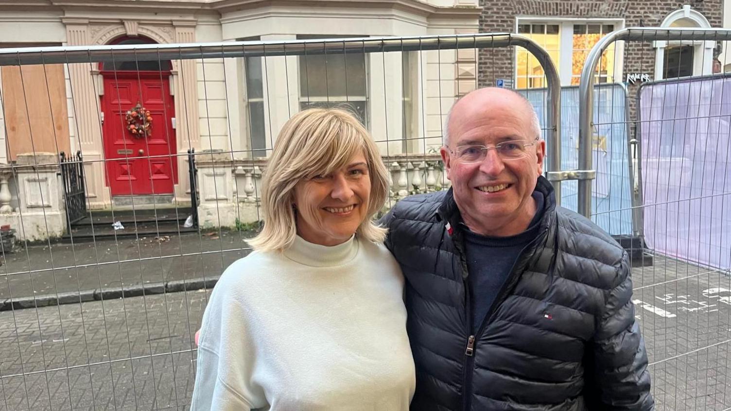 Noel and Maria Mason are standing side by side in front of metal railings near a building with a bright red door and boarded up windows. Noal is wearing a navy coat and glasses, while Maria has wavy blonde hair and is wearing a white jumper.