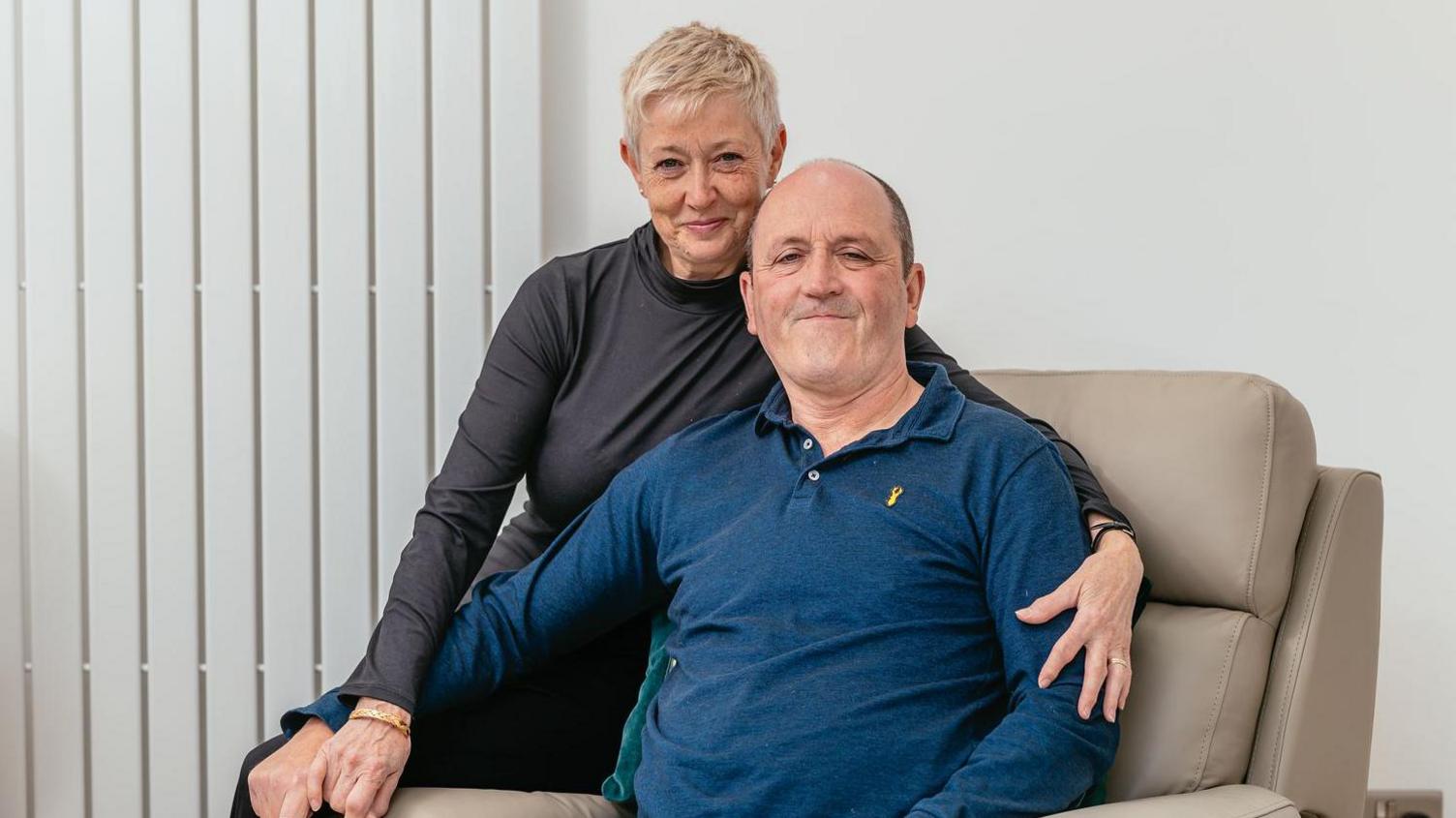 Mike Thomas, who has a bald head and is wearing a blue polo top, sat in a cream-coloured armchair and smiling while looking at the camera. Jayne who has short blonde hair and is wearing a black, long-sleeved turtle neck t shirt is sat behind him, on one of the arms of the chair, and has her arms round him