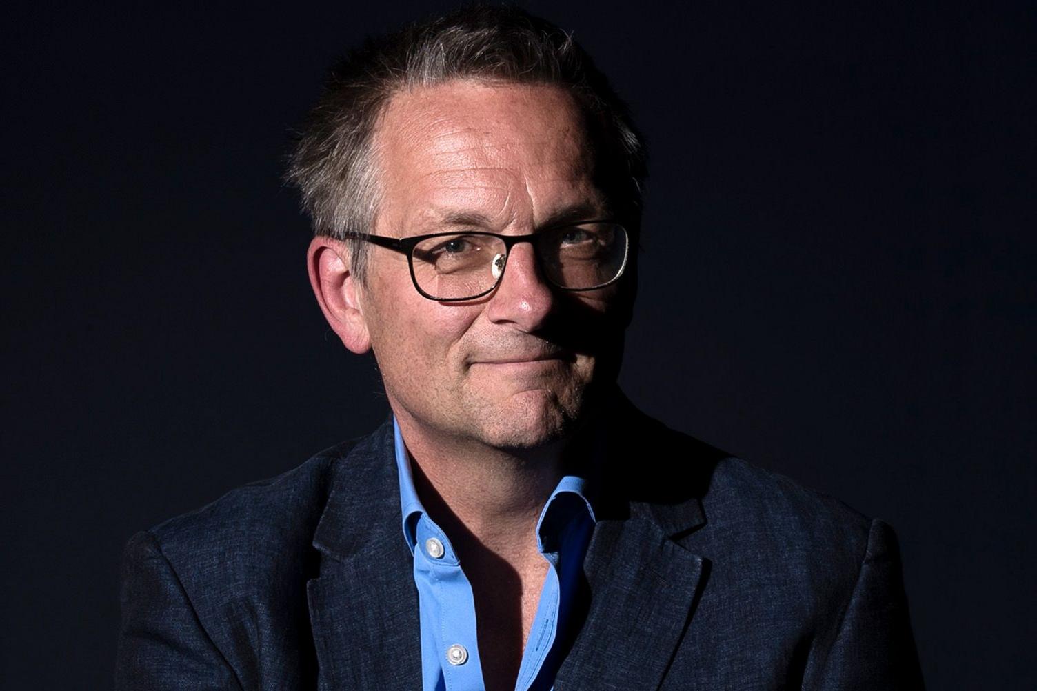 Publicity shot of Michael Mosley against a black background. He has glasses, slightly receding greyish hair, and is smiling as he looks to the camera. He wears a dark jacket and a cobalt blue shirt with the top two buttons undone.