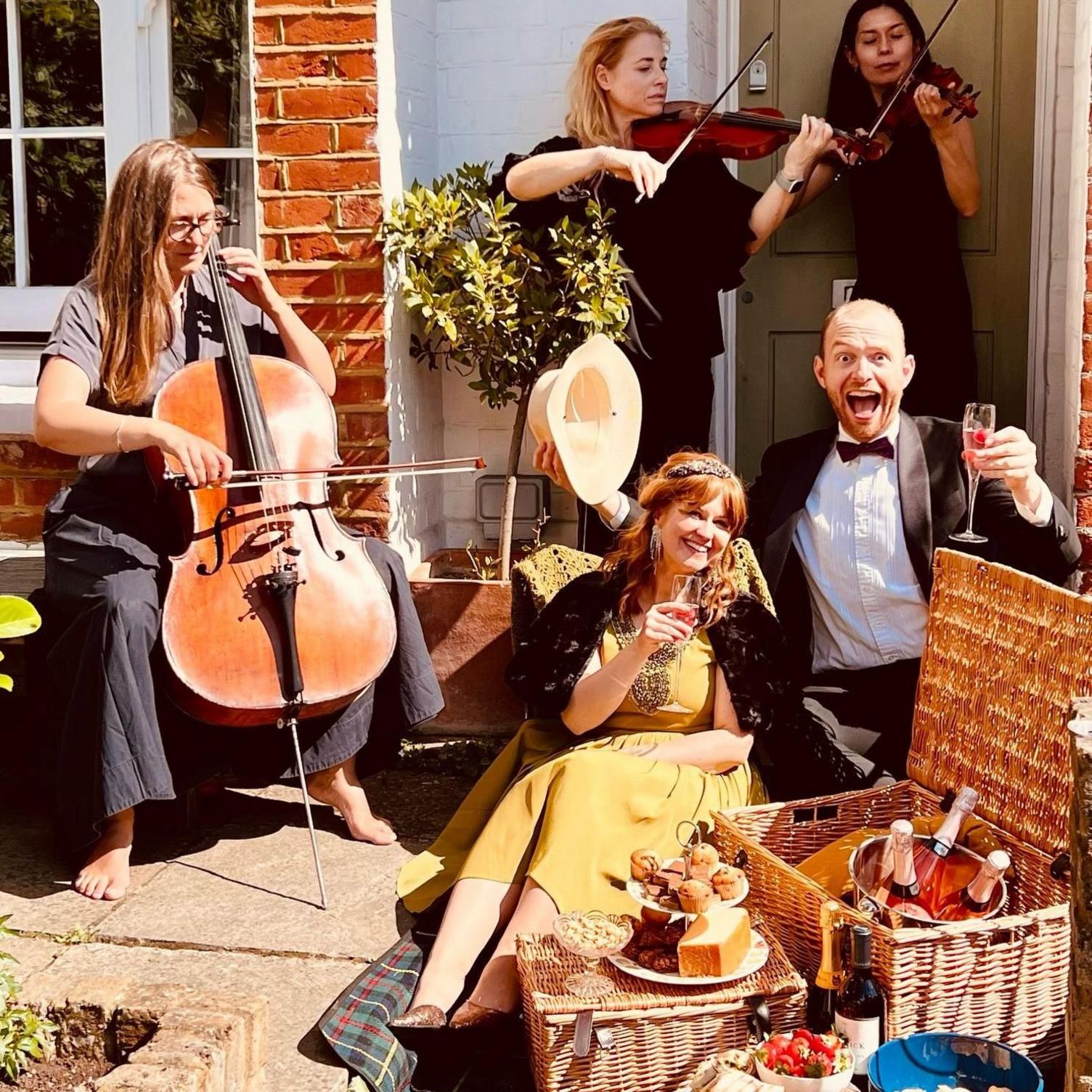 Tom, in a dinner suit and bow tie, and Beth, in a gold dress, sit on a picnic blanket outside their house with glasses of fizz. Behind them, three string players perform while a hamper overflows with food and wine.