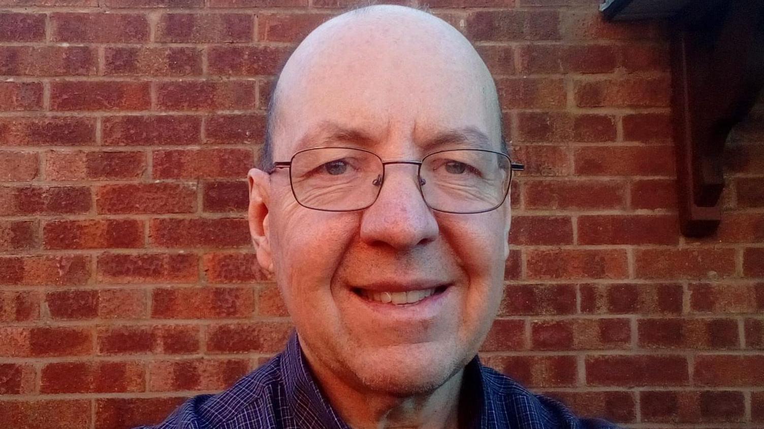 A selfie of a smiling Andrew Bullock standing in front of a brick wall, outside. He is 61, bald and wearing glasses.