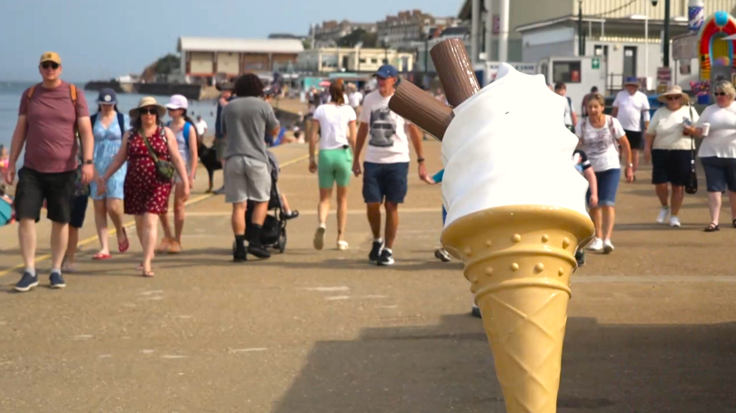 We can see a busy seaside promenade, with lots of people walking on it. At the front of the picture is a large plastic ice cream cone, with ice cream and flakes in it.