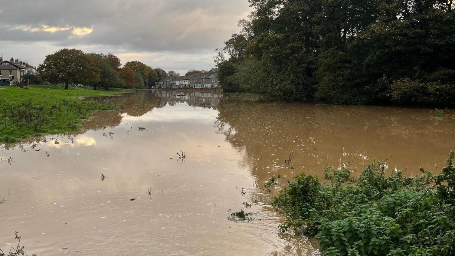 A large stream of brown muddy water is flooding an area of pastureland. The water stretches out to the houses in the distance. On the left is a grassy slope of pasture with flood water washing across. 