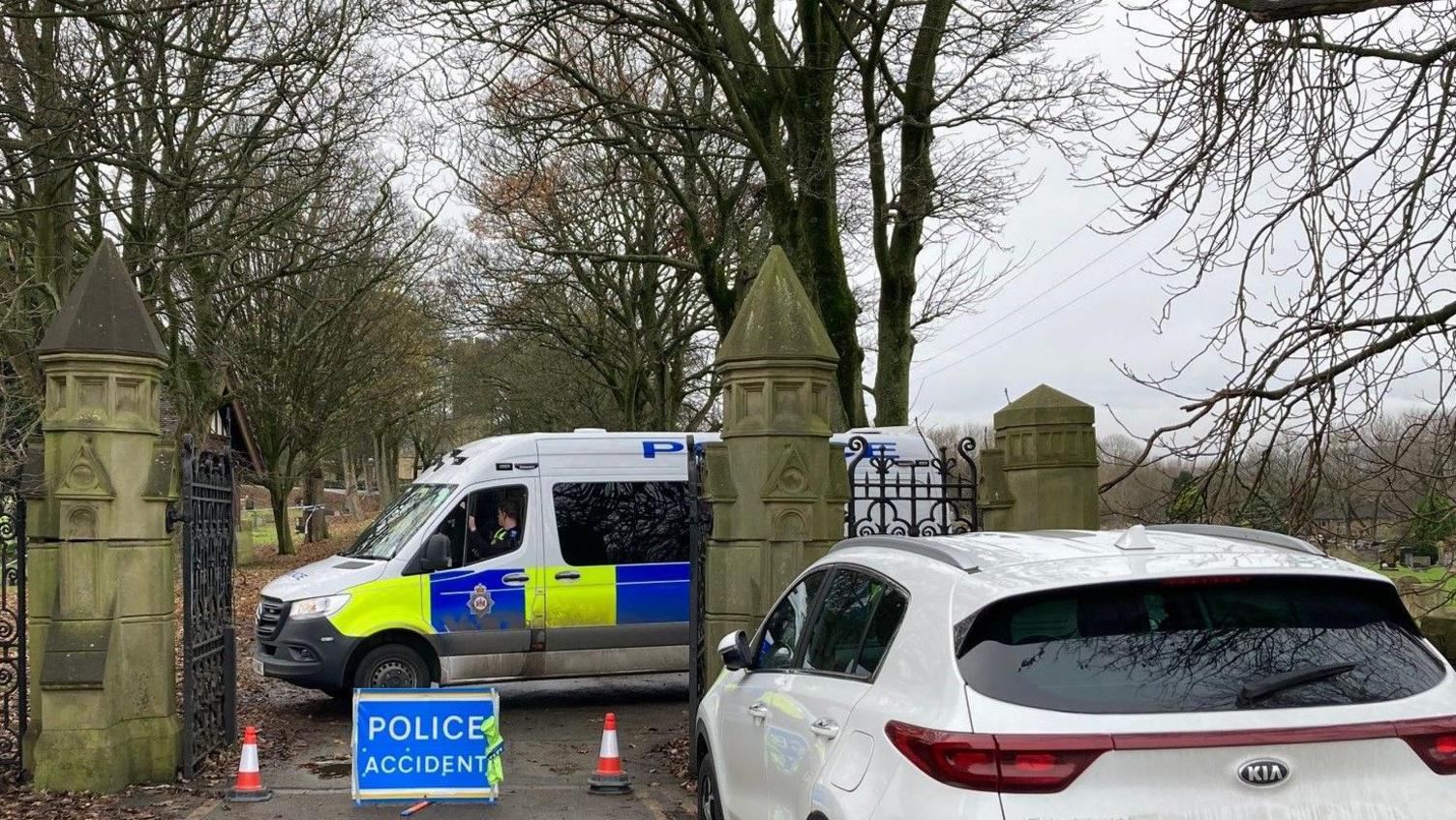 A blue and white sign saying 'police accident' and two red and white traffic cones blocking off an entrance to Scholemoor Cemetery with a police van parked behind the cordon.  