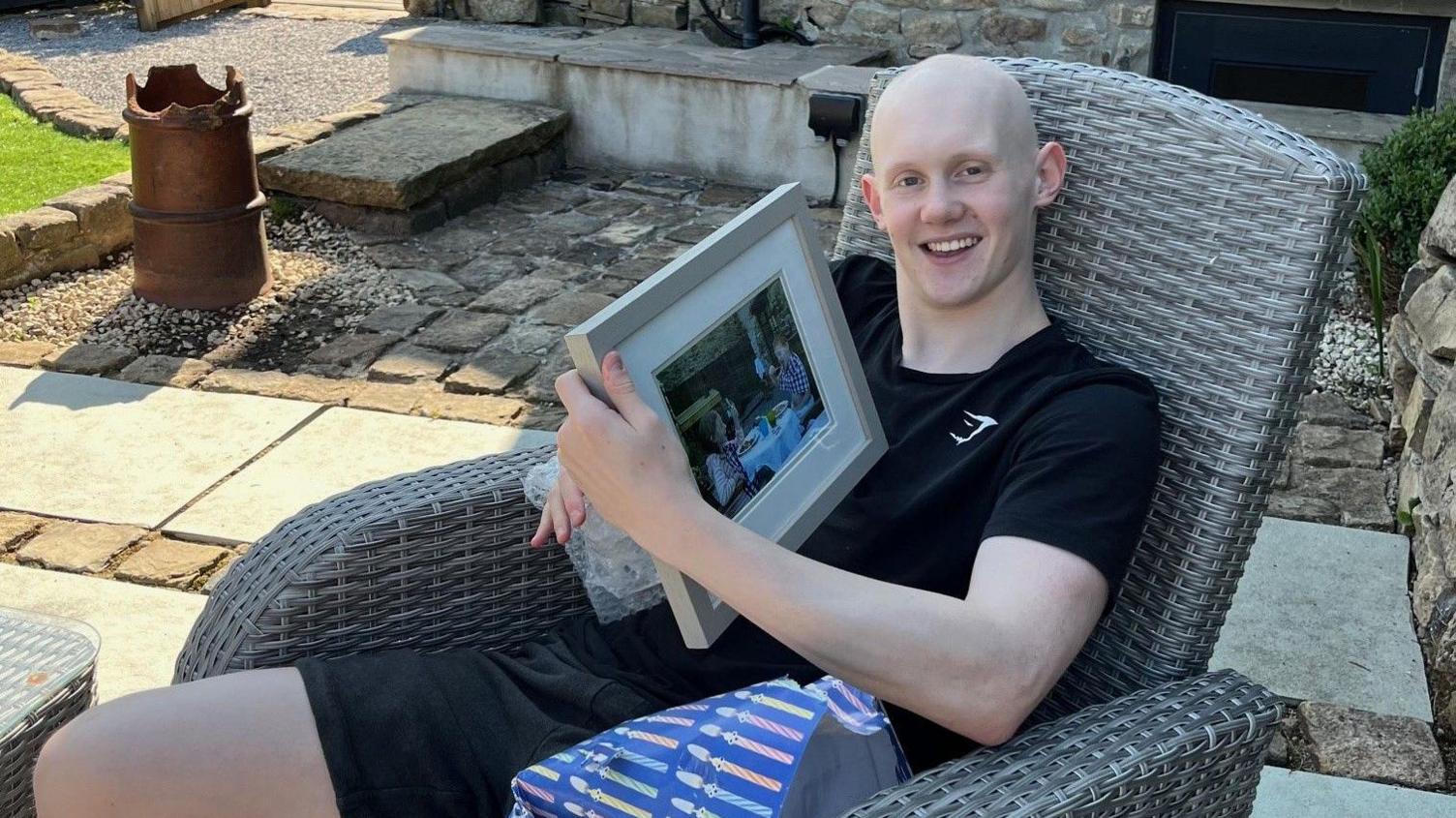 A boy is sitting in a garden chair holding a present which he has unwrapped. The present is a photo or picture in a frame.