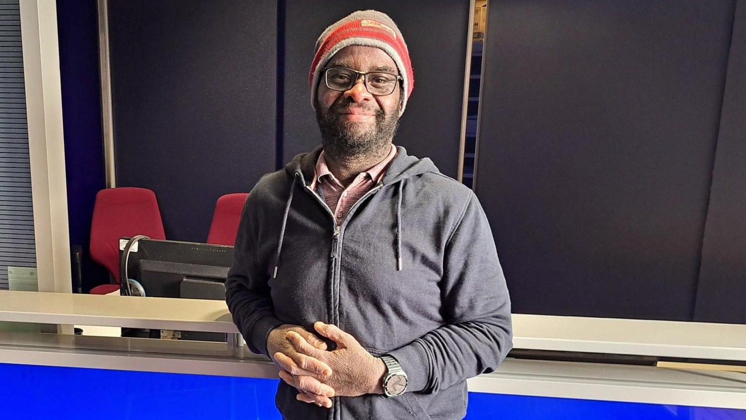 A man wearing a blue hoodie and red hat with his hands clasped together. 