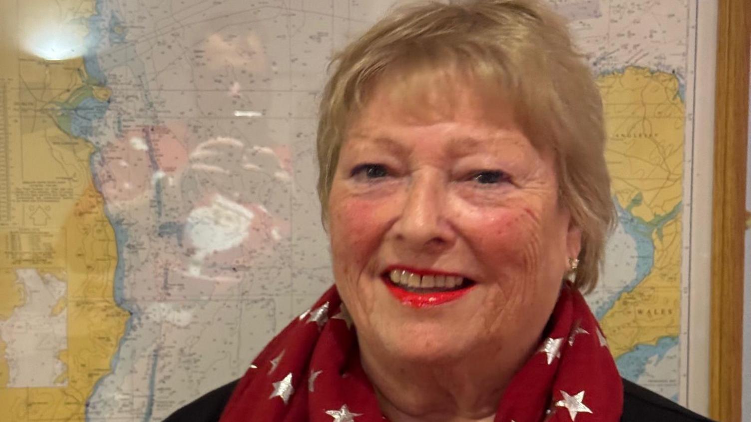 Gilli Cowley, pictured with short blonde hair, wearing red lipstick and a red scarf with a map in the background