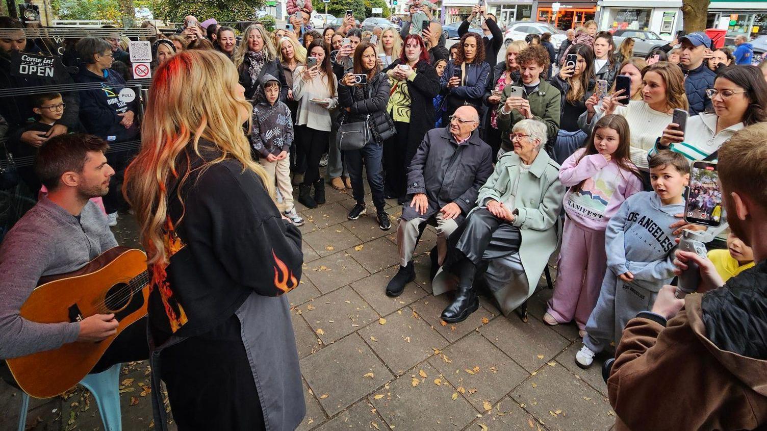 A large crowd standing on the pavement watching Ella Henderson sing along with a man playing acoustic guitar.