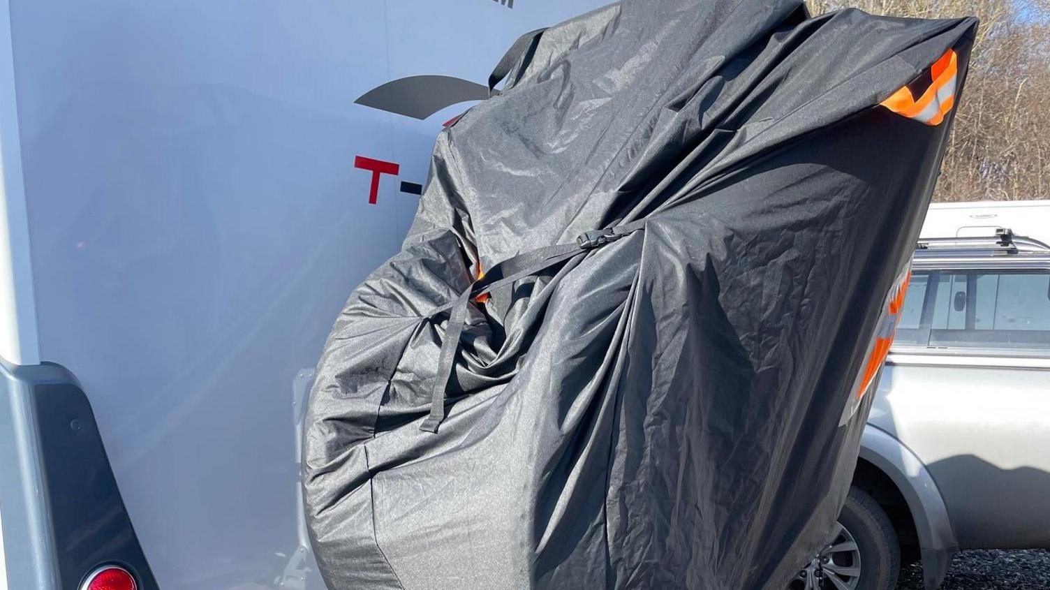 A tightly-packed cover on the back of a van. It is pictured outside on a driveway or road.