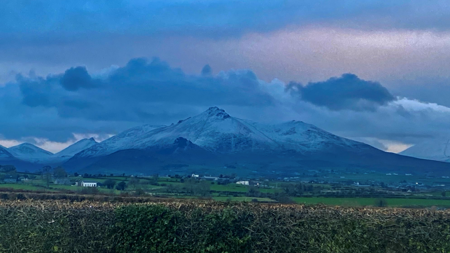 Several mountains, their peaks topped with snow, can be seen in the distance against a cloudy sky background. In the foreground can be seen green fields and hedges.