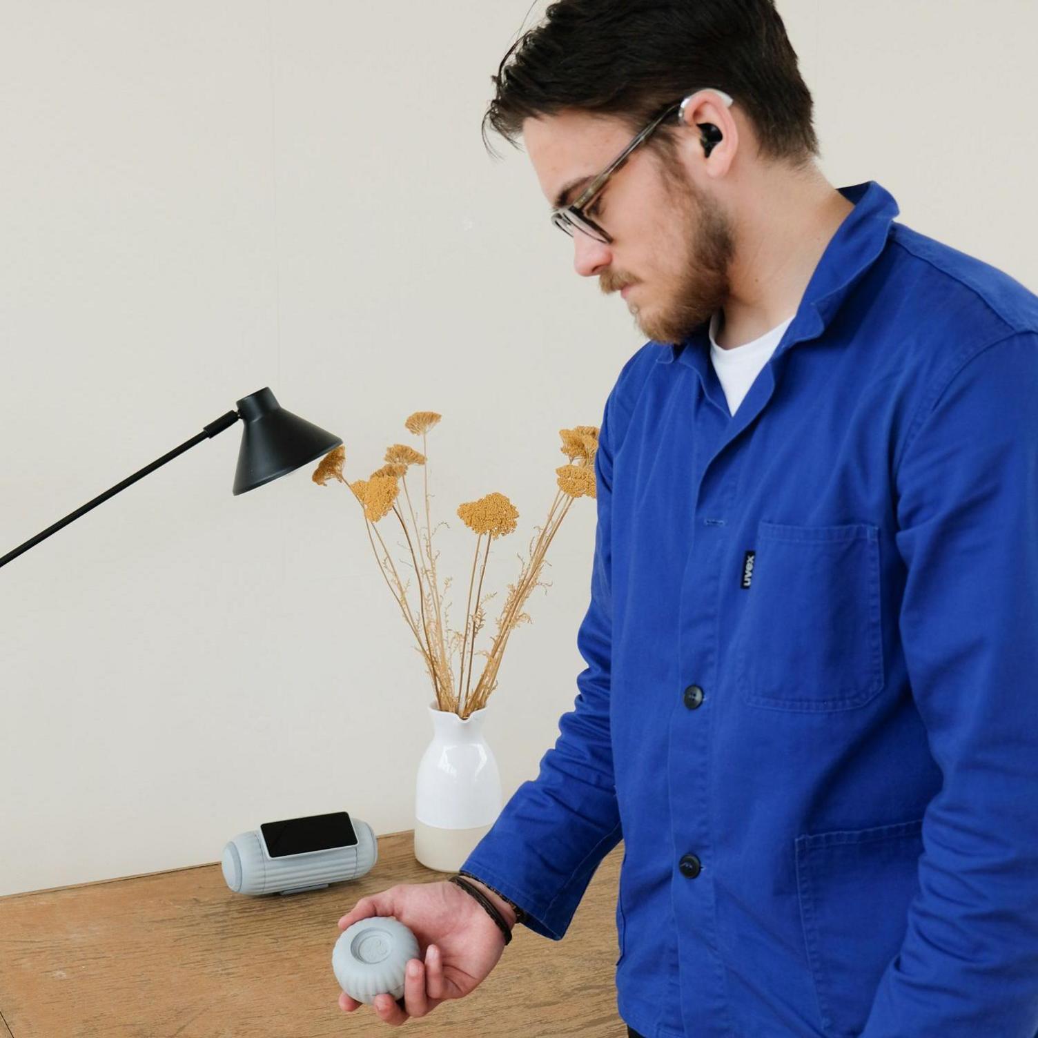 Sam Russell is pictured with both elements of the smart speaker. He is wearing a bright blue shirt with a white T-shirt underneath and has dark hair, dark facial hair and wears glasses and hearing aids