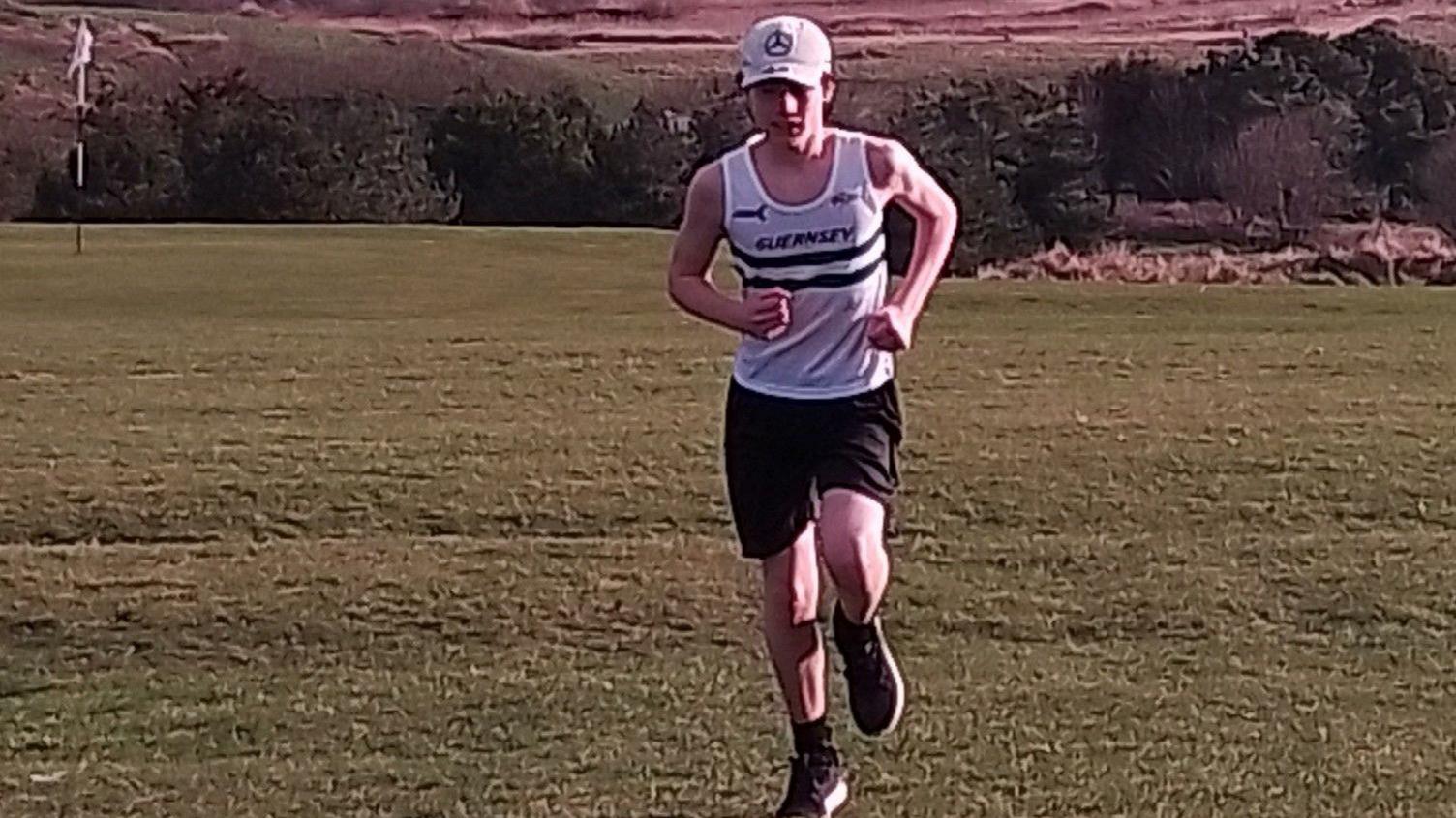 George running over what looks like a golf course towards the camera. He is wearing a white vest, black shorts and a white cap.