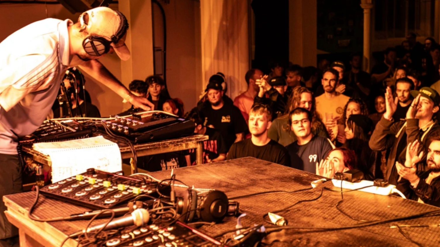 DJ on stage wearing a white t-shirt and cream cap. A large audience is watch. They are clapping and dancing to the performance. The stage has a lot of sound equipment on there not being used. 