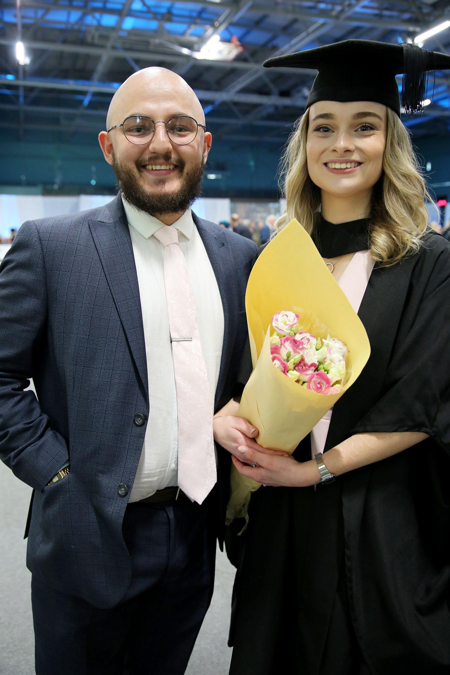 A man standing on the left. he is bald, with circle framed glasses and a brown beard and moustache. He is smiling standing with his hands in his pockets. He is wearing a dark blue suit, white shirt and a pink tie, with a silver tie pin.
A woman standing next to him on the right is wearing a graduation cloak and hat. She is blonde and also smiling. She is wearing a pink dress and a silver watch. She is holding a bouquet of pink and white roses wrapped in yellow paper