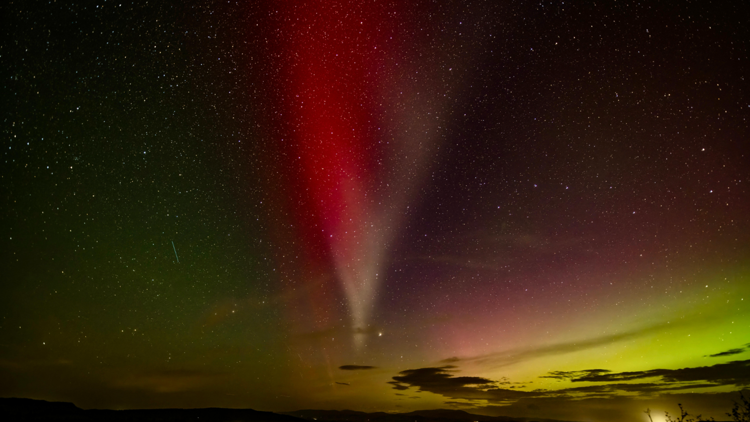 Night sky with stars and a ribbon of white and red light across the sky. In the background, hints of purple and green of the Northern Lights