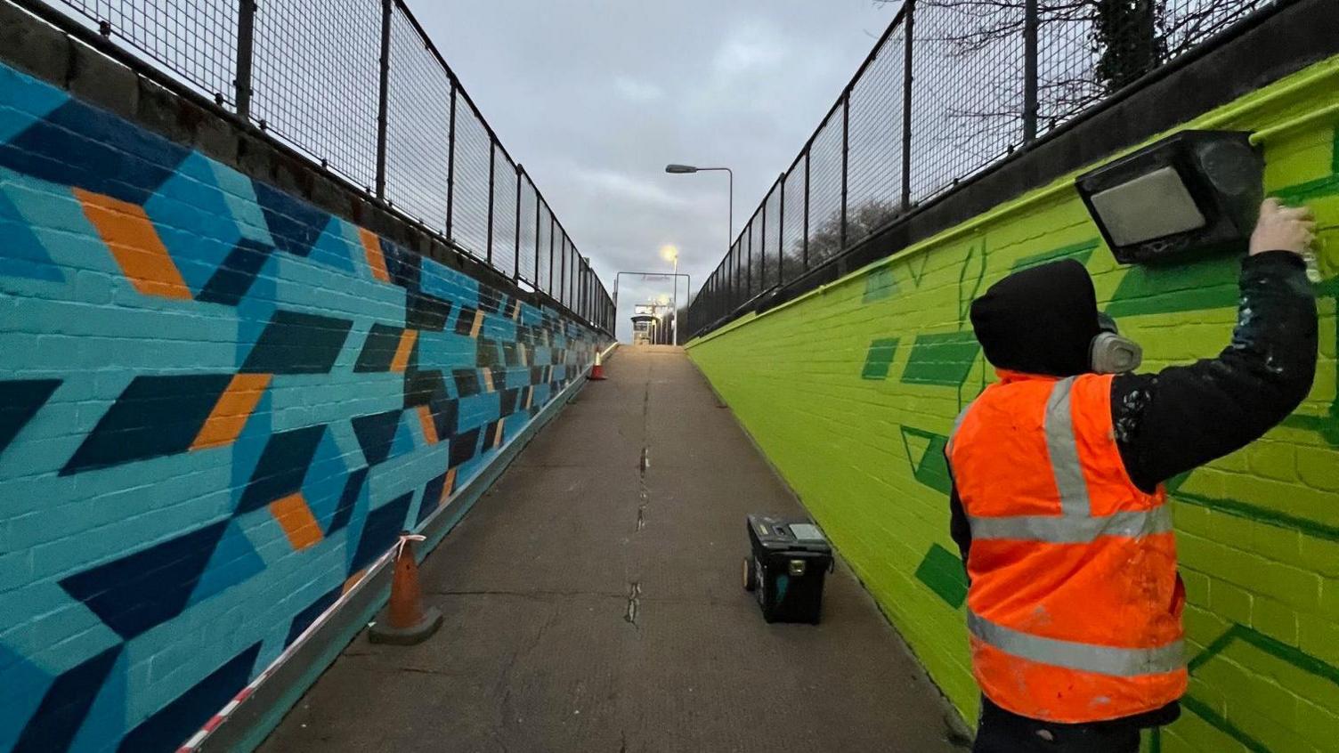 The image shows an underpass at Bedminster train station, taken from the bottom to the top of the underpass where the station platform is. On the right side of the image, a man is spray painting the wall that is covered in different shades of green in a pattern. He wears a florescent jacket and a black hoodie covers his face. On the opposite wall, a pattern is painted in various shades of blue and orange. 