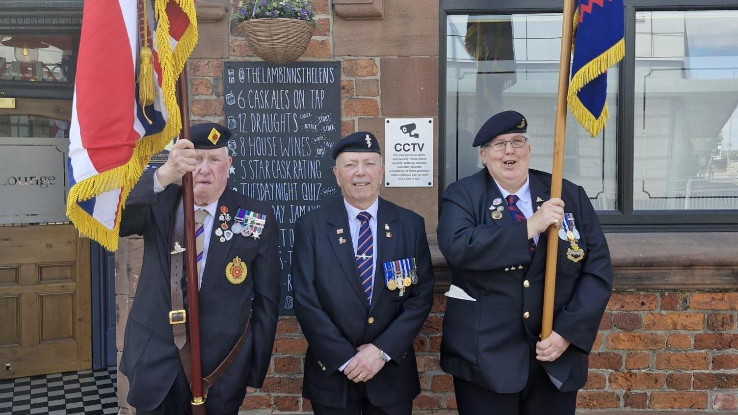 Three veterans in dark coloured suits with a number of medals and wearing berets with military pin badges stand outside the pub. Two of them hold military flags.