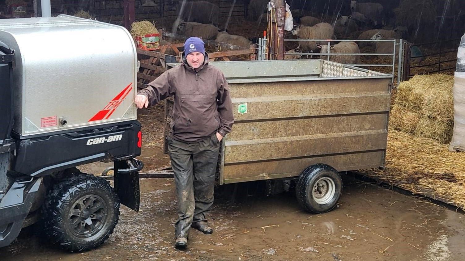 Mark Mather stands in front of a trailer in the pouring rain with sheep behind him.