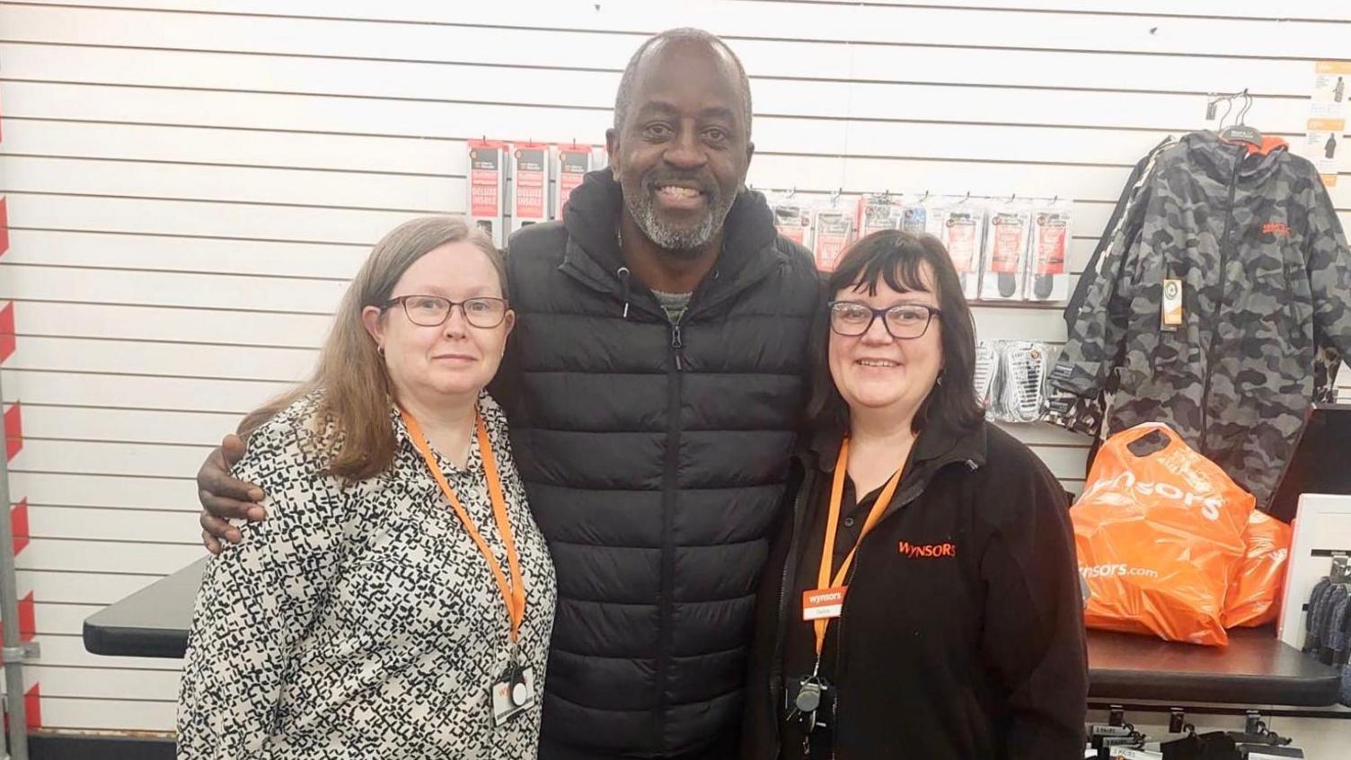 A man and two women hug in a shoe shop. The woman on the left has glasses, long brown hair, a black and white blouse and on orange lanyard. The woman on the right has the same lanyard, a black fleece with an orange logo, glasses, short black hair and glasses. The man between them has a grey goatee beard and a black puffer jacket. Behind them is a black shop counter with two orange bags on it. Shoes soles and camouflage jackets hang from the walls which have horizontal white stripes.