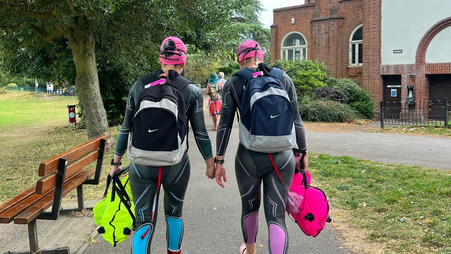 Georgia and Melissa Laurie walking away from the camera wearing their wetsuits, waterproof rucksack and bright pink swim hats touching each other's hands ahead of the race