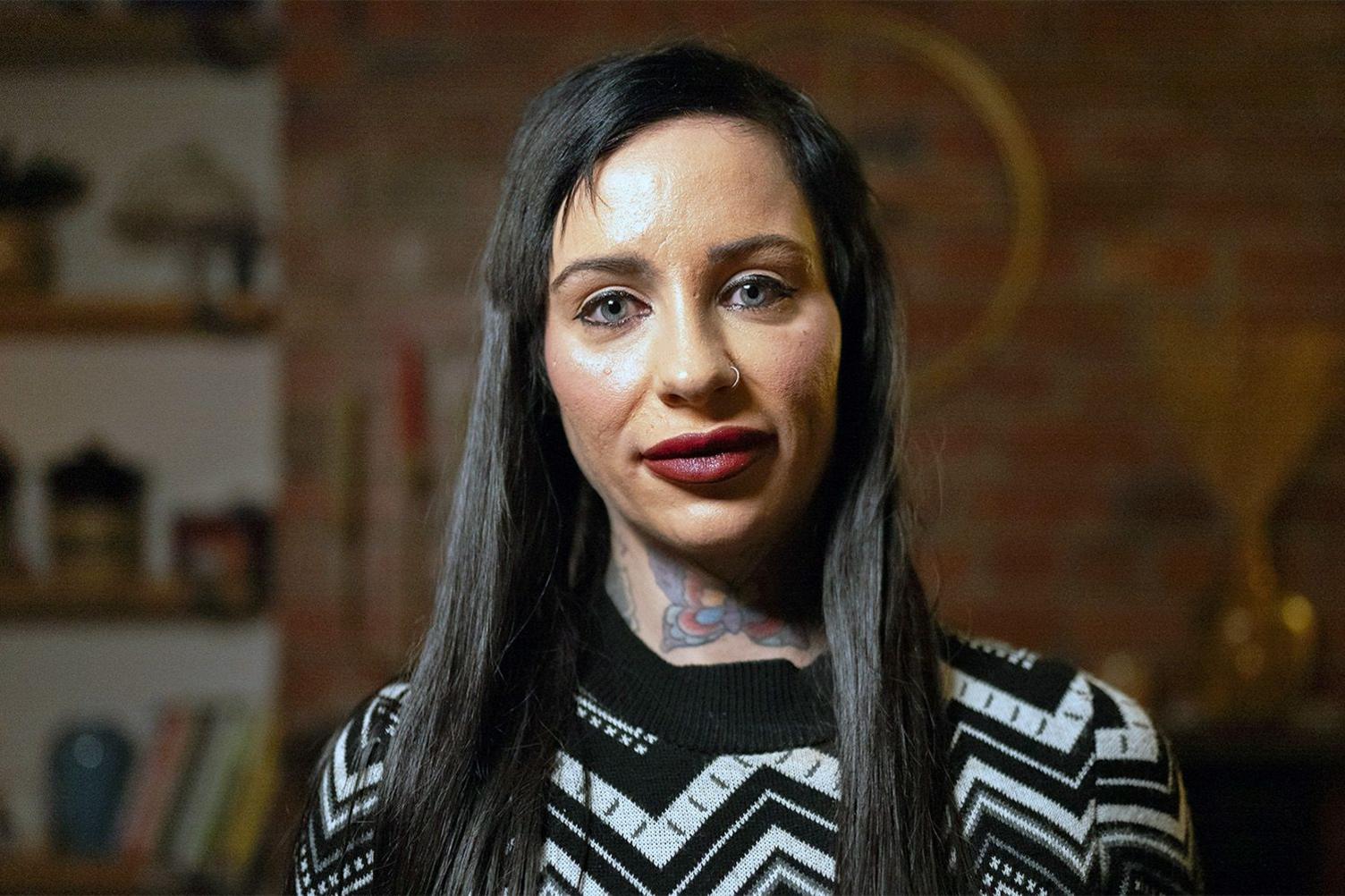 Portrait shot of Lisa Elliot in a living room. She has long, straight, dark brown hair and blue eyes. She has a tattoo of a butterfly on her neck and a nose ring, and is wearing a black and white striped jumper.