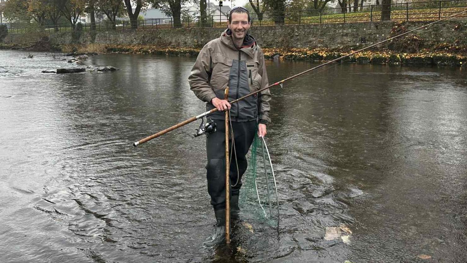 Fisherman Kieron Banks stands with a rod and a net in the River Kent. He is wearing fishing waders and a large green fishing coat. It is a wet day with the trees on the river bank losing their leaves.