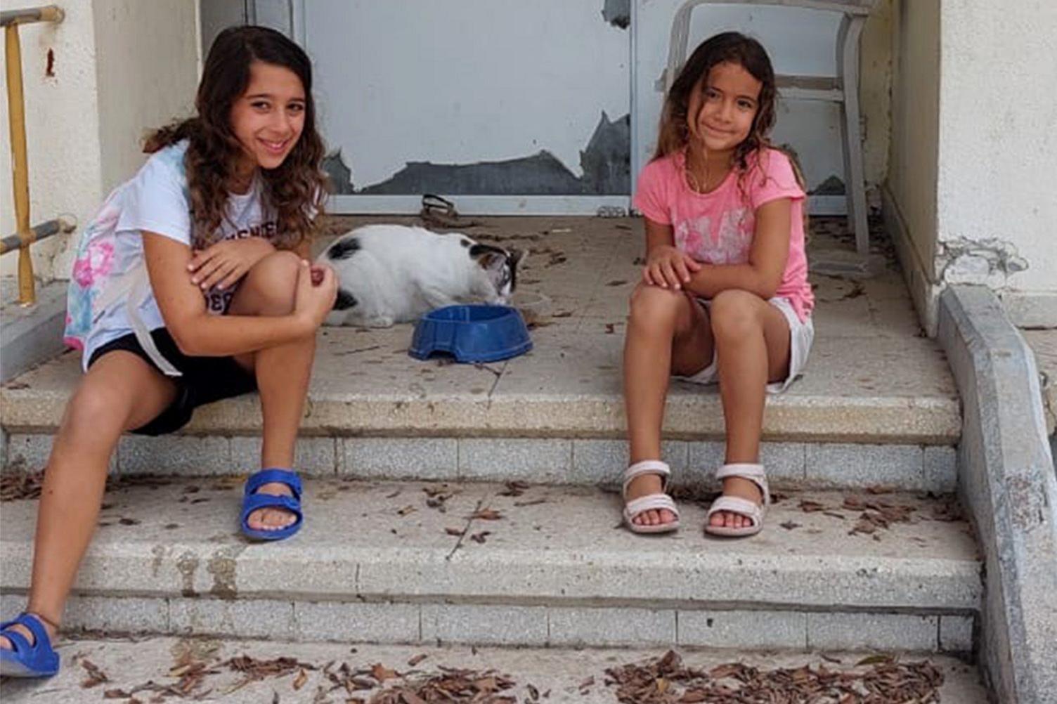 Shir's daughters, both with long dark hair and smiling, sit on the steps of their home - next to them a cat eats out of a cat bowl