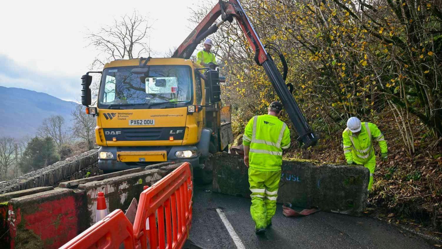Three workmen use a crane attached to a lorry to move large concrete blocks into the middle of the road.