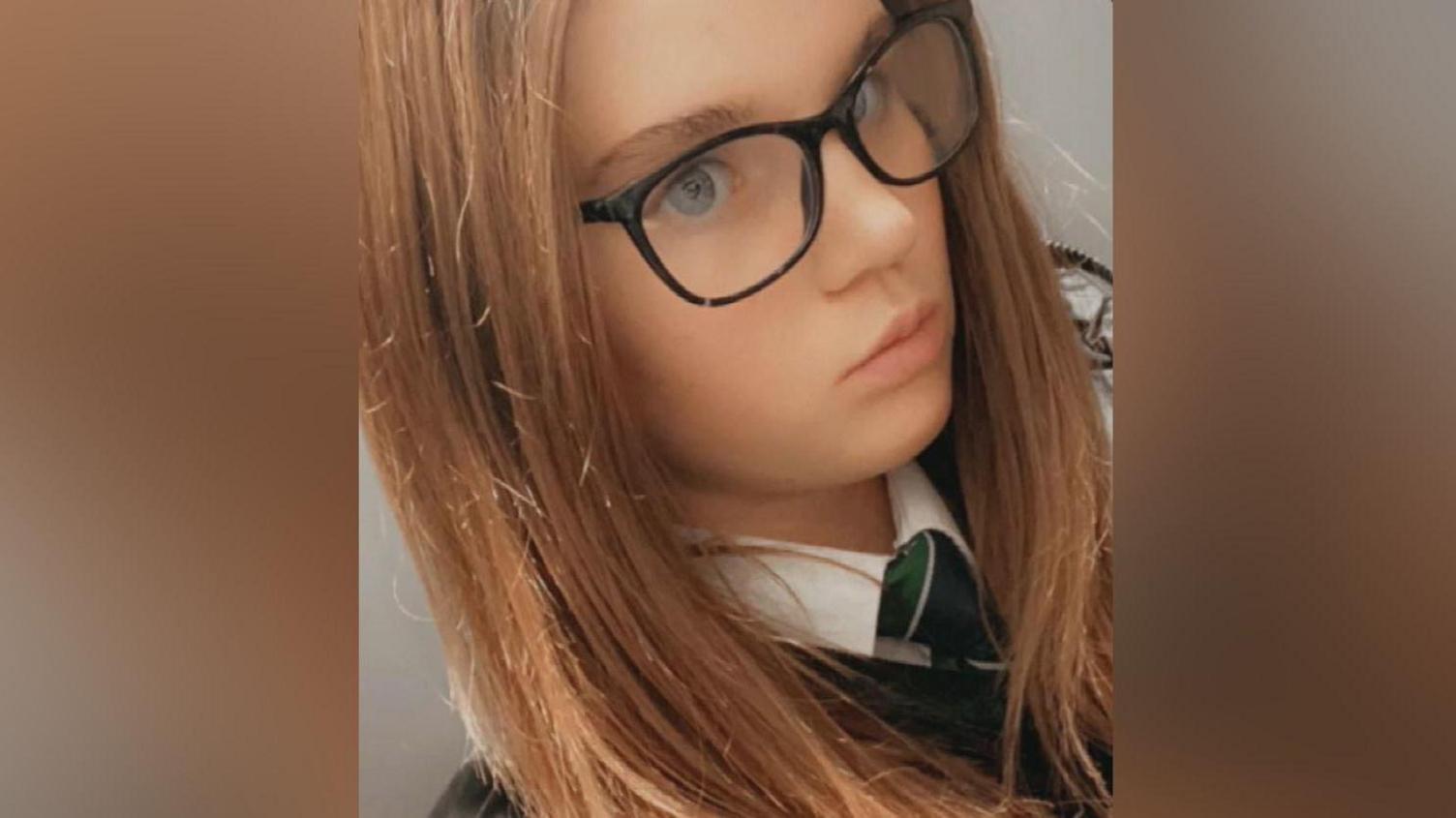 Holly Newton, a young girl with long dark hair wearing a white school shirt with a green tie and glasses takes a selfie.