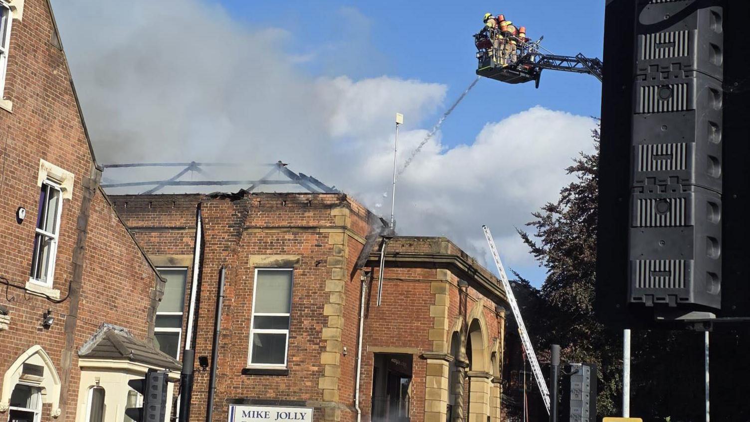 Firefighters stand on a cherry picker over a smoking building with a damaged roof