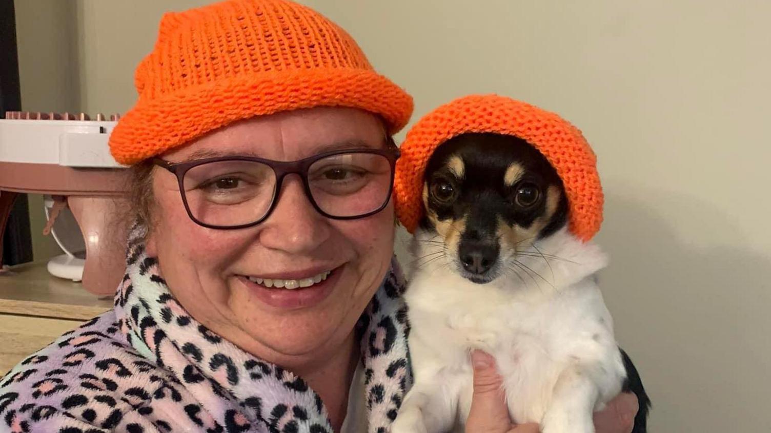 Denise Appleton and her dog. Denise and the dog are wearing orange knitted hats. Denise is wearing a leopard-print dressing gown. 