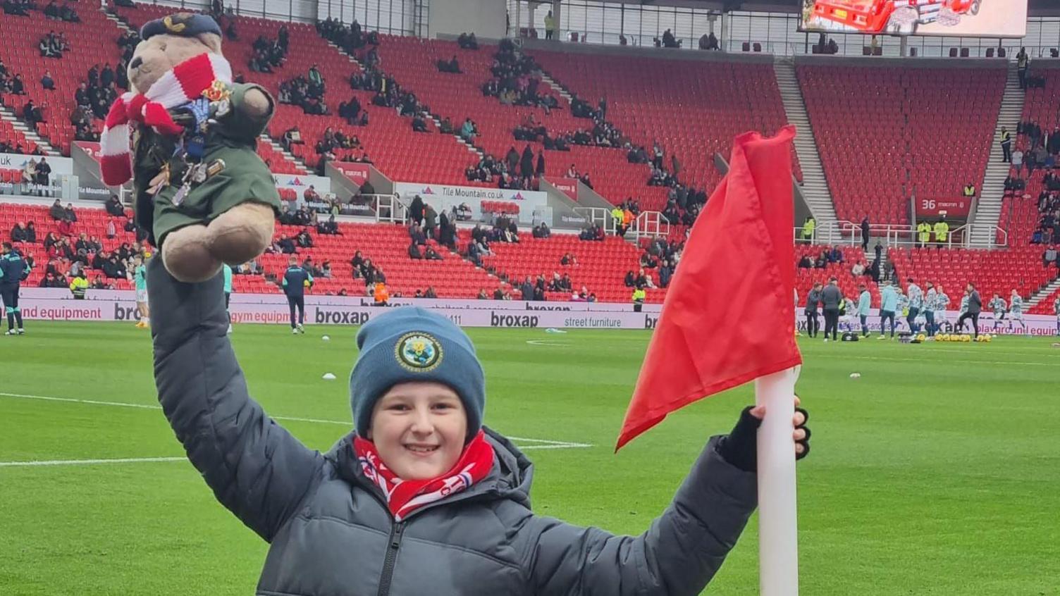 A child holds a teddy bear aloft by the corner flag of a football stadium.