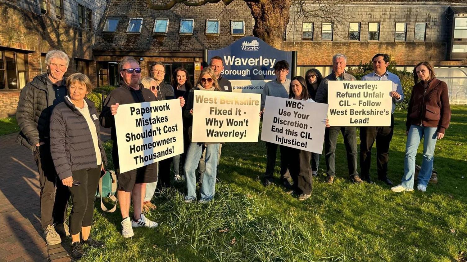 A group of people stand in front of a Waverley Borough Council sign on a grassy area with building in the background. They are holding signs which read "paperwork mistakes shouldn't cost homeowners thousands" and "WBC: Use your discretion - end this CIL injustice".