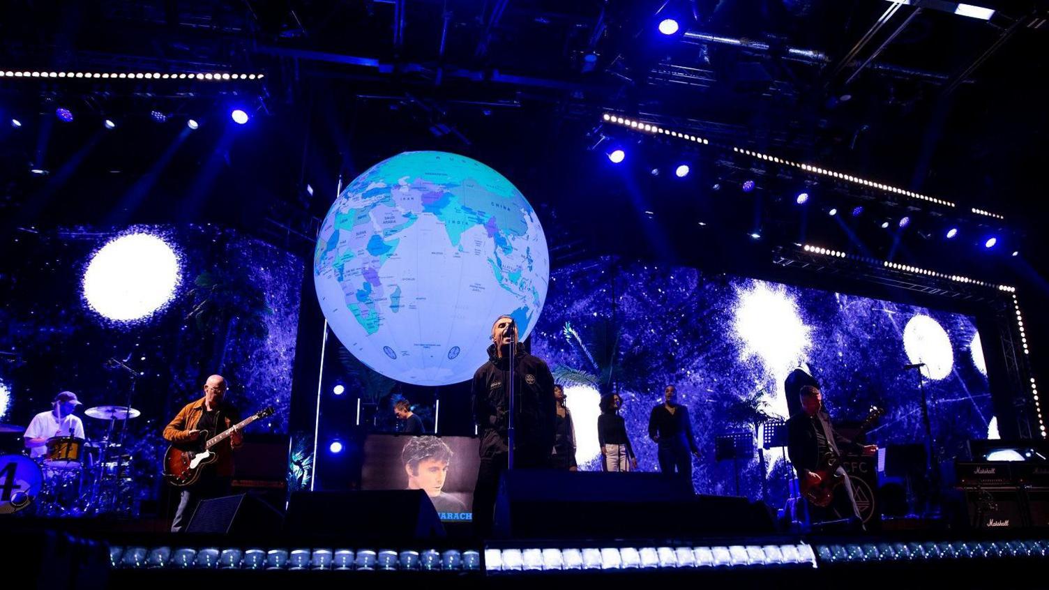 Liam Gallagher on stage with musicians and backing singers at the O2 Arena in London. The backdrop is dark blue with a number of large white, fuzzy edged circles and the stage props include a giant globe suspended above them