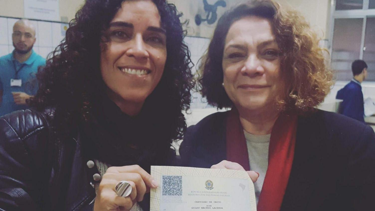 Two women smiling, looking at a camera, and holding up a document