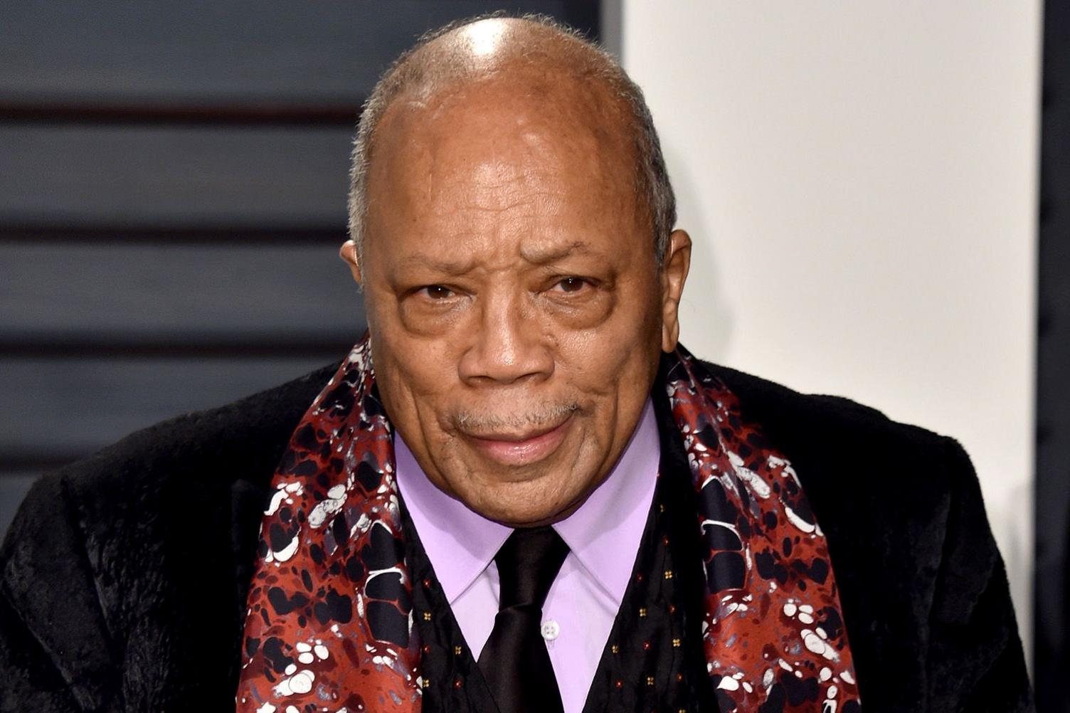 News photo of Quincy Jones, smartly dressed in a dark suit, purple shirt and black tie, accessorised with a brown abstract-pattern silk scarf. He is quite elderly in this photo, with no hair and a thin moustache.