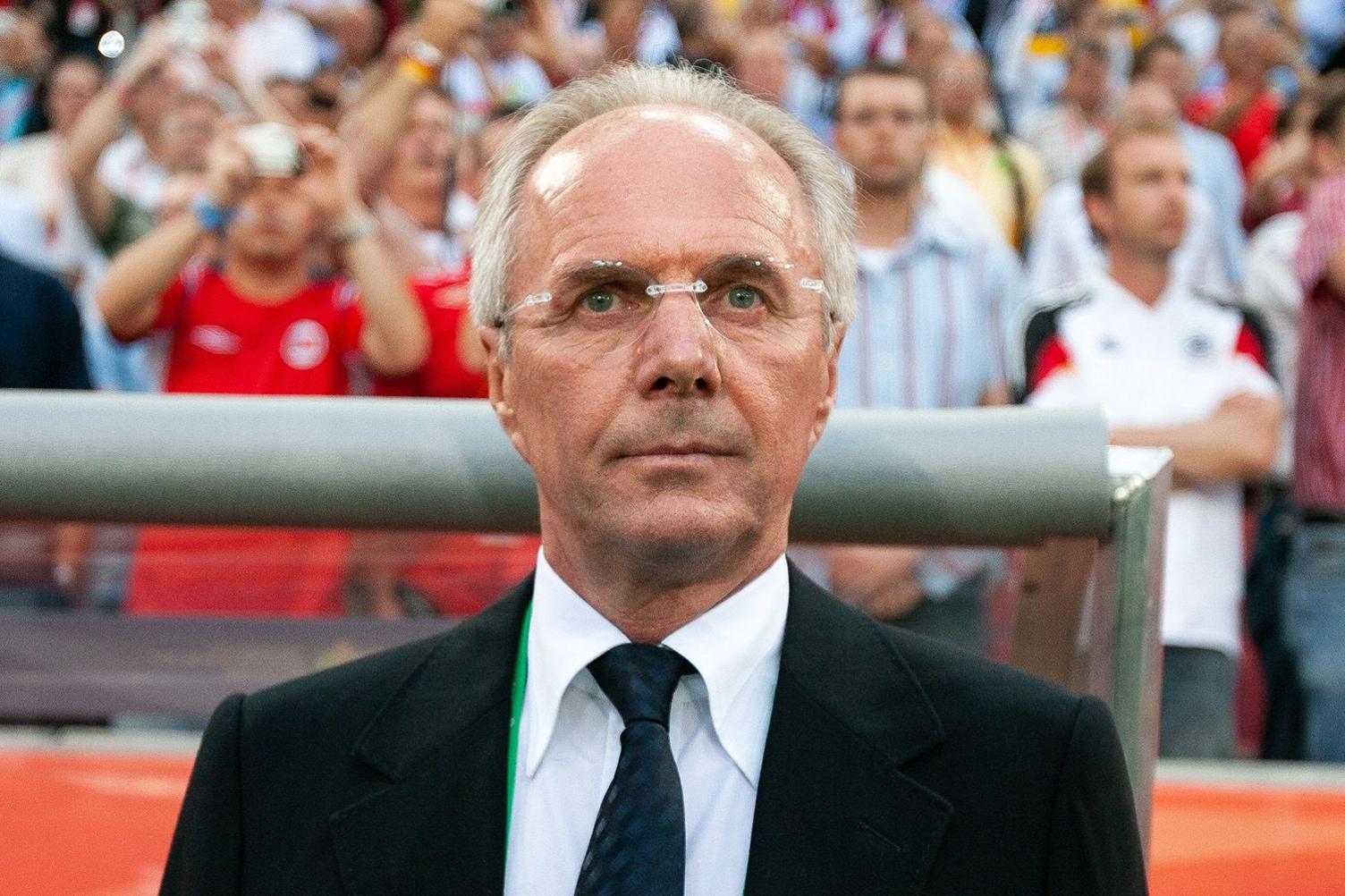 Sven-Goran Eriksson looks impassively beyond the camera in front of a crowd of spectators in England replica shirts. He has receding grey hair and wears frameless spectacles, a black suit jacket, white shirt and dark tie.