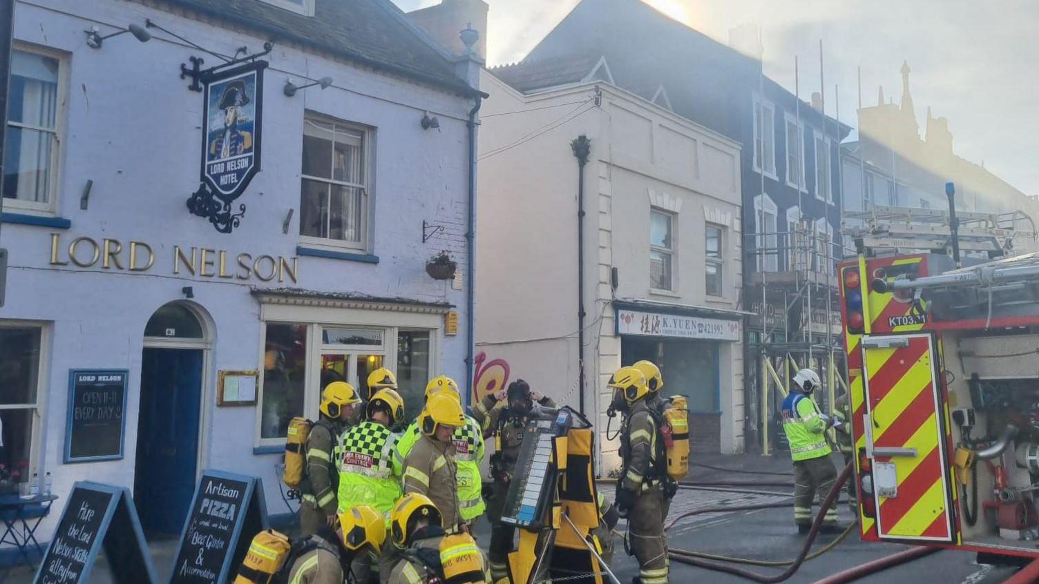 A collection of firefighters with equipment on and stood next to a fire engine. There is smoke rising from a building nearby on East Street.