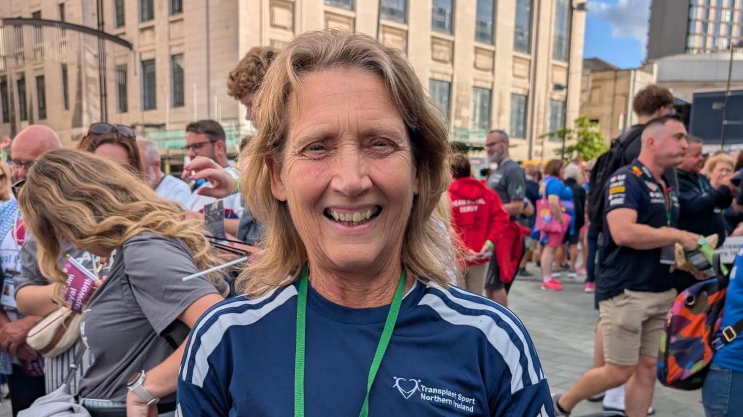 Kay Hack has long blonde hair and is wearing a navy T-shirt and green lanyard. She is standing in front of a large crowd of people.