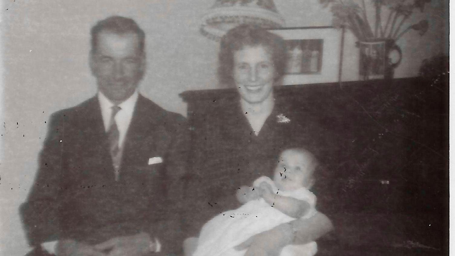 A black-and-white photograph of Bob and Margaret Armstrong. Bob is wearing a suit and tie with a handkerchief in his top pocket, while Margaret has a dress with a broach on the lapel and is holding a baby