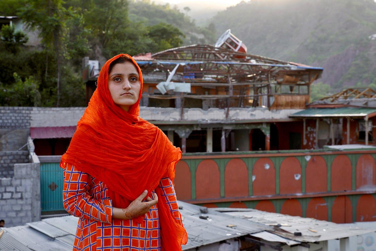 Nimra, 16, wearing an orange headscarf and a patterned outfit stands in front of the damaged mosque, mountains visible in the distance.