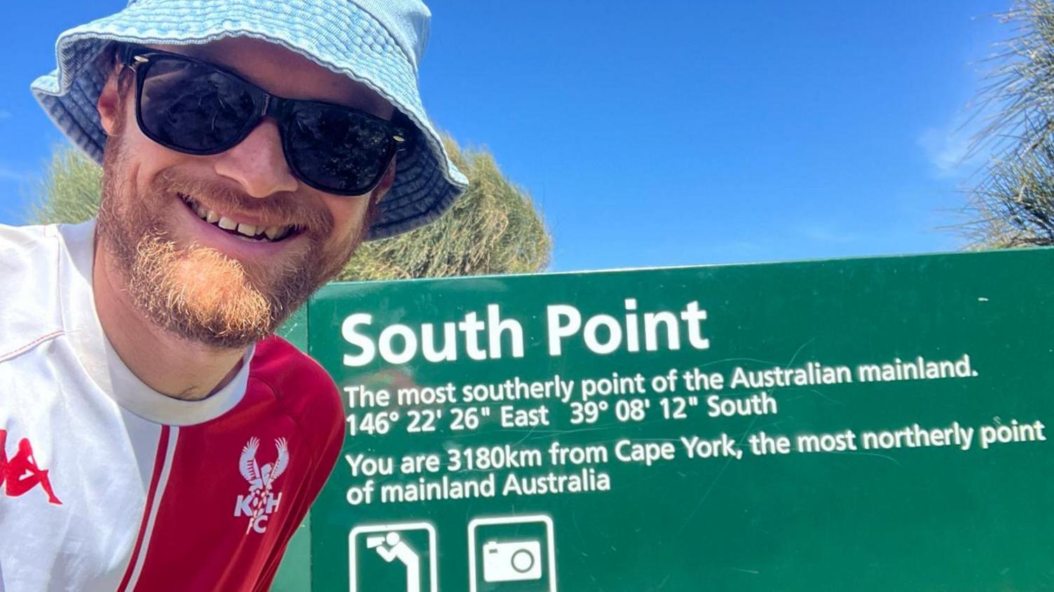 A man with a gingery beard wearing sunglasses, a pale blue bucket hat and a half-red, half-white football top is smiling at the camera below a clear blue sky. Next to him is a sign with some map coordinates which reads: "South Point - the most southerly point of the Australian mainland. You are 3180km from Cape York, the most northerly point of mainland Australia."