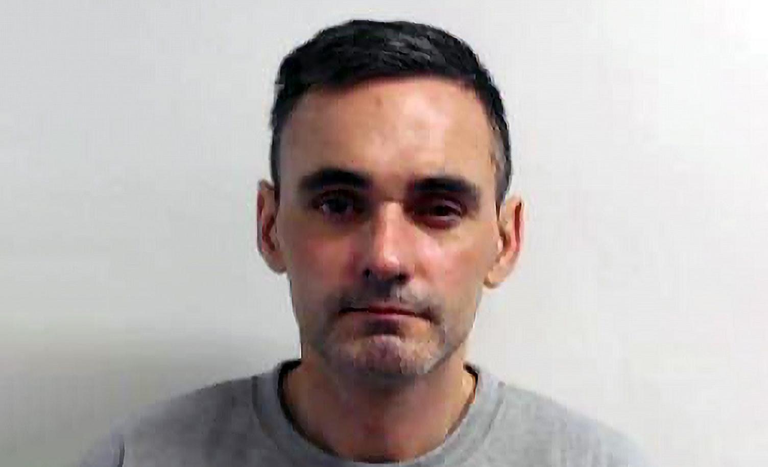 A police mugshot of a middle age man with short dark hair and grey stubble in a grey sweater