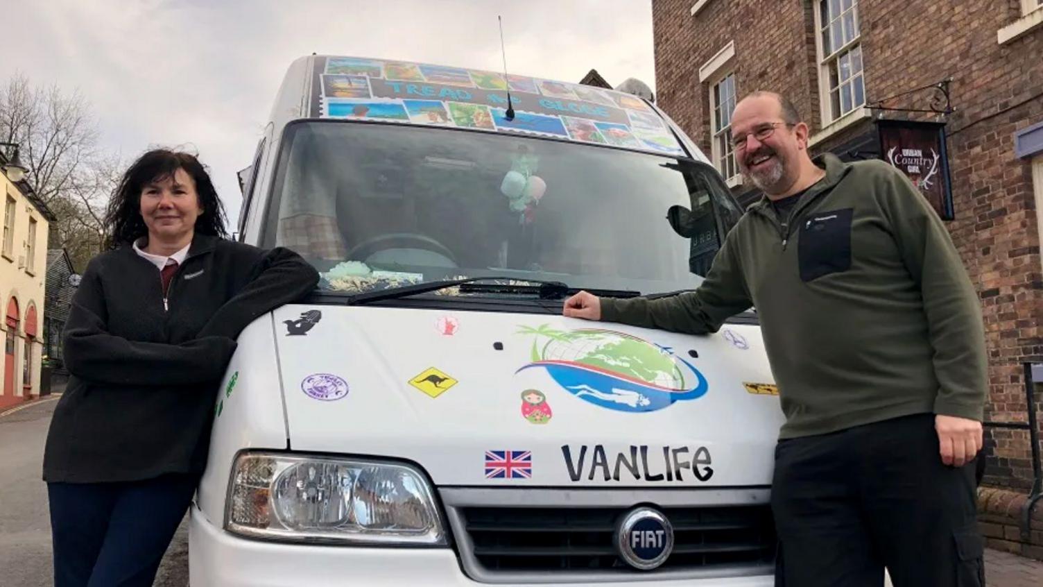 A woman in a black zip collar jumper and a man in a green zip collar jumper lean against a white camper van, which is covered in stickers and graphics and says Vanlife quite clearly on the front