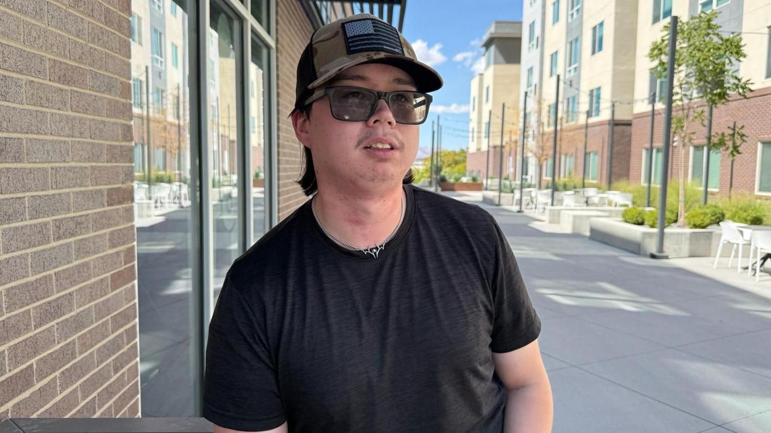 Scott Sperry is seen wearing a black T-shirt, a camo hat and sunglasses. He is outside in a courtyard of a student housing complex.