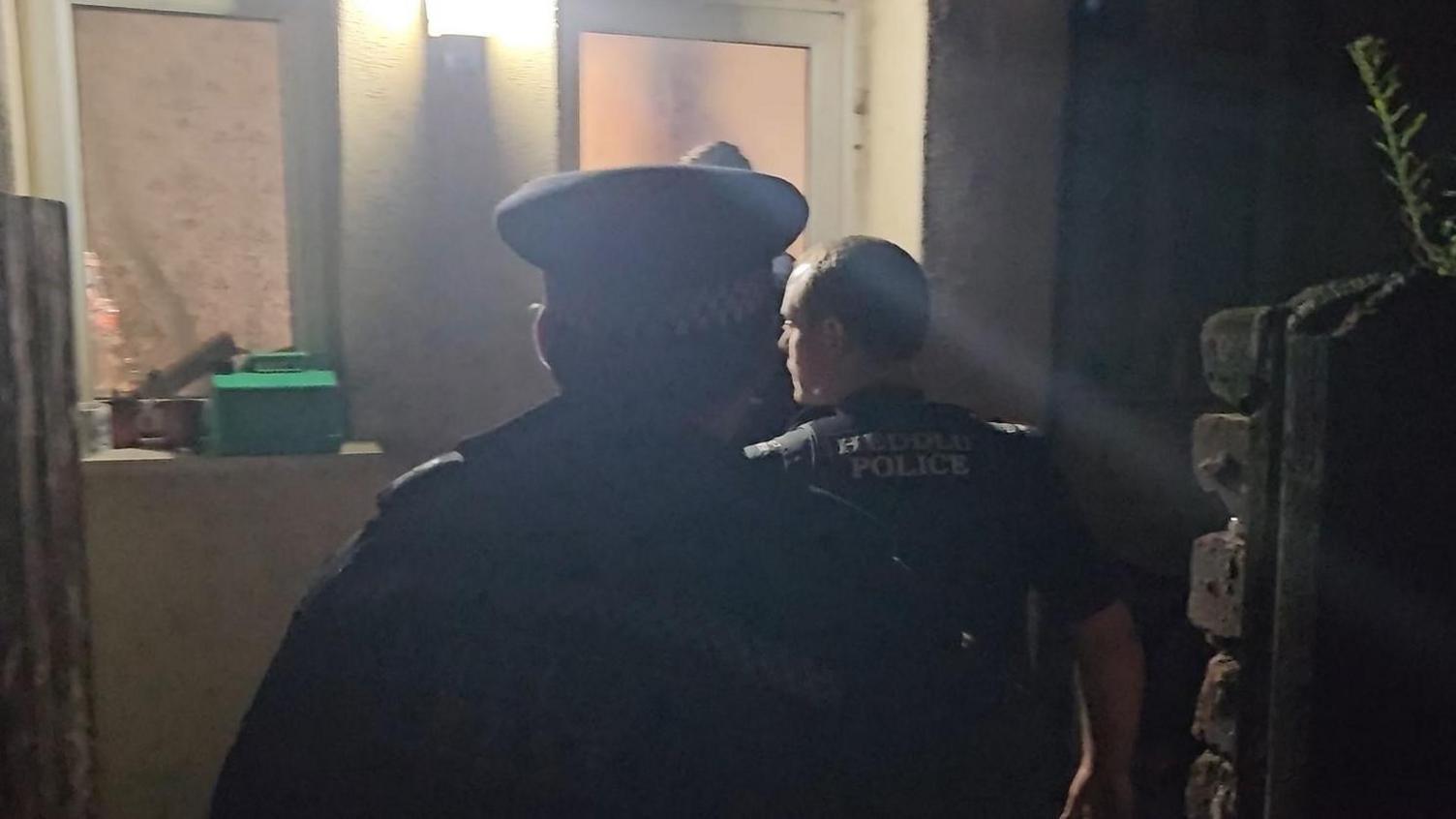 Three silhouetted police officers approach the white back door of a house in Rhyl under cover of darkness