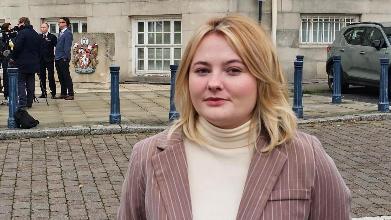 A woman in a pink blazer with pinstripes over a white jumper. She is stood outside a large stone building and has a stern expression.